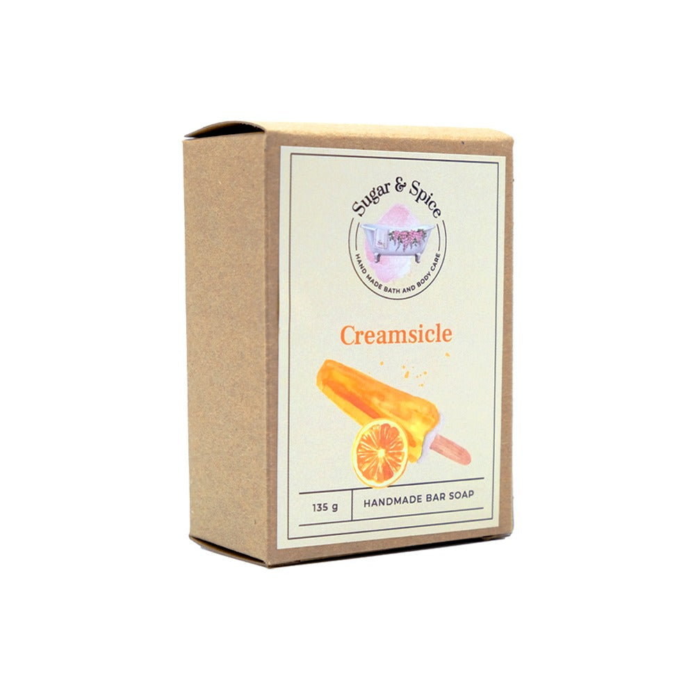 A packaged bar of Creamsicle handmade soap with the brand name Sugar & Spice visible on the label. The packaging is primarily beige with an illustration of an orange slice, indicating the scent or ingredient of orange in the soap.