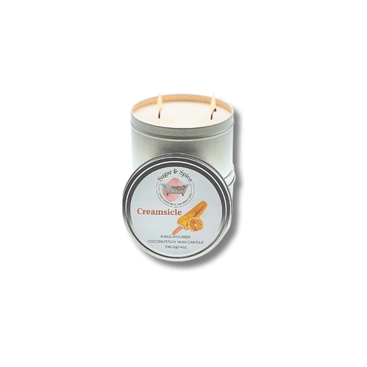 A silver-colored, metal tin container with a lid, containing a two-wick Creamsicle-scented coconut wax candle. The label on the tin has orange and white colors and text.